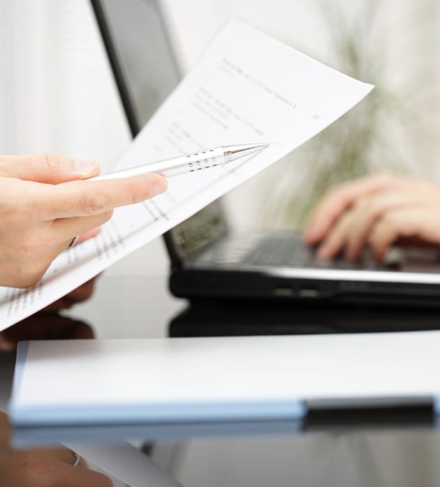 close up of hands holding a paper and a pen pointing to blured type on the page, person typing in the background
