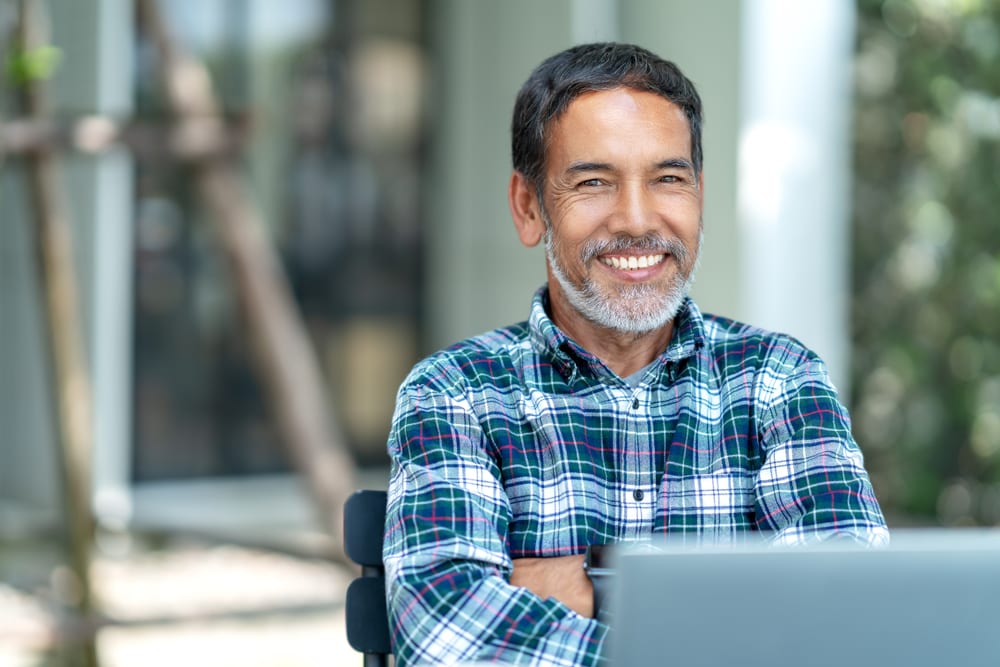 Happy mature man with white, grey stylish short beard looking at camera over his laptop screen outdoors