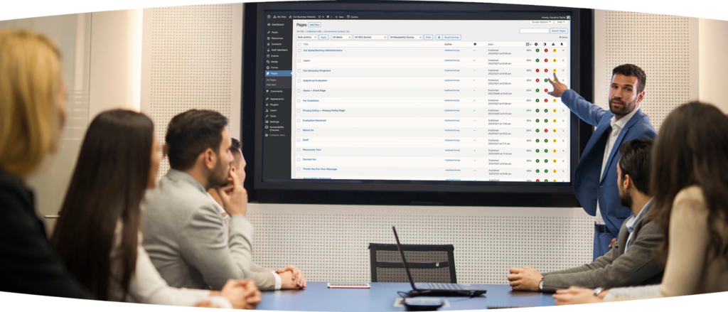 Six men and women sitting around a conference table and looking as another man stands next to a large monitor pointing to an Accessibility Checker pages report on it.