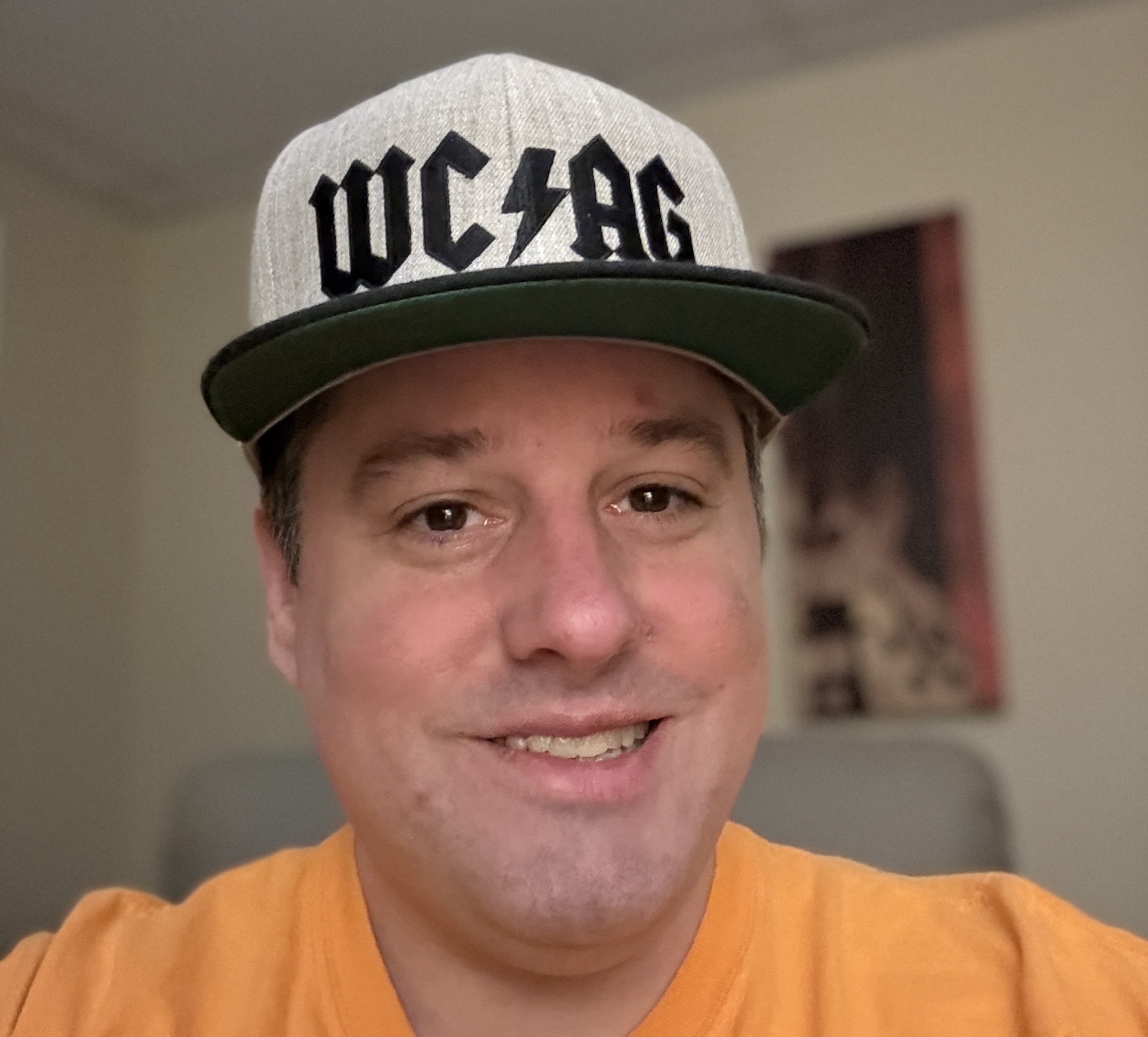Steve Jones smiling at the camera wearing a grey “WCAG” baseball cap and orange shirt, seated indoors.