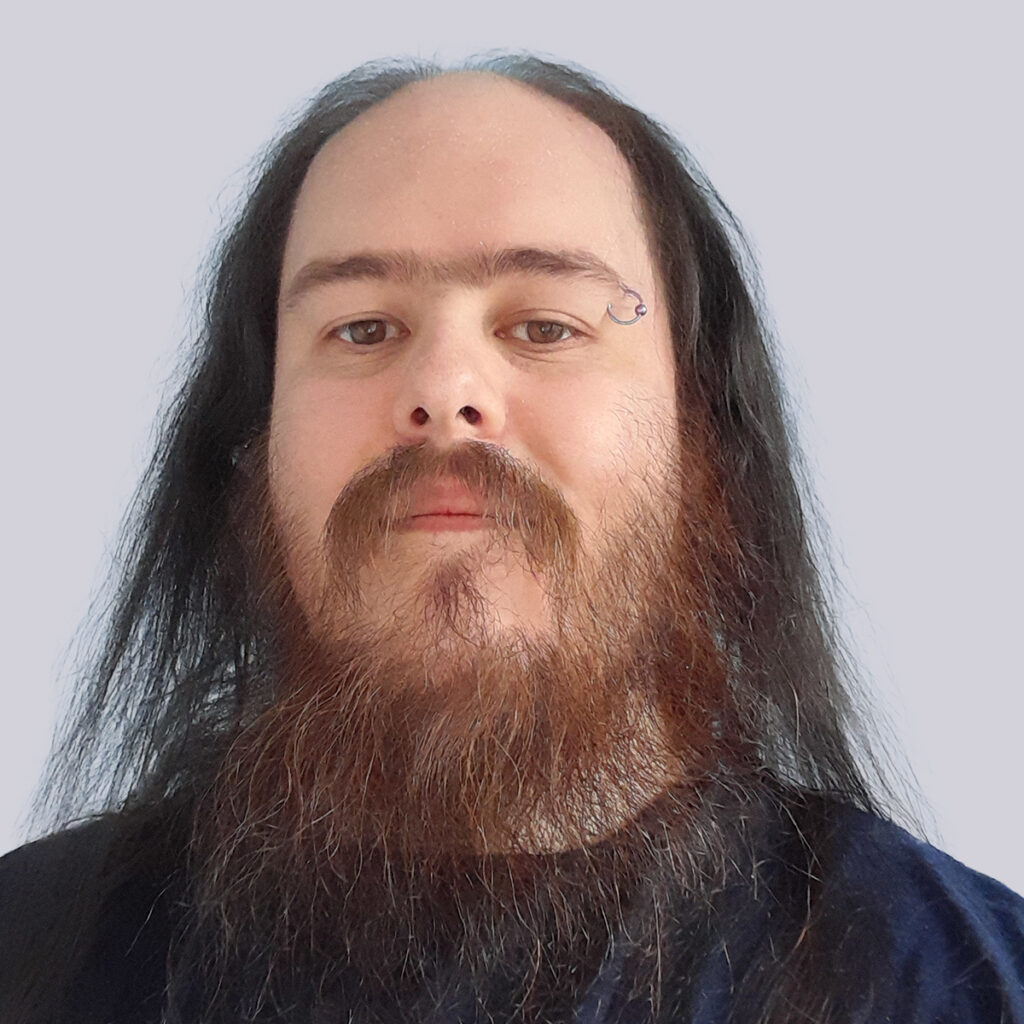 A person with long dark hair and a thick, reddish-brown beard wearing a dark-colored shirt is photographed against a plain light gray background.