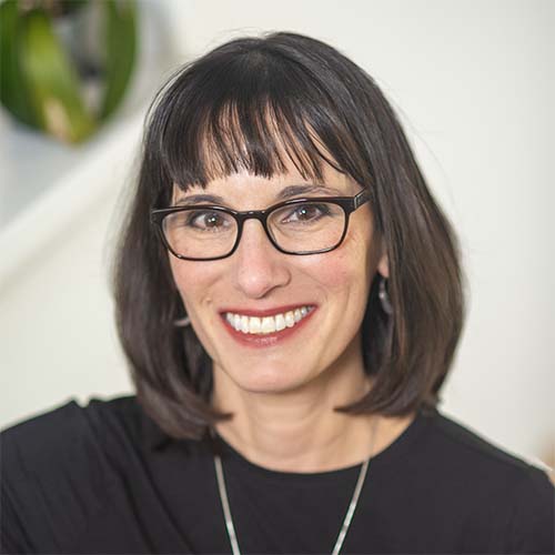 A middle-aged Caucasian woman with shoulder-length brown hair and bangs wears a black top and glasses.
