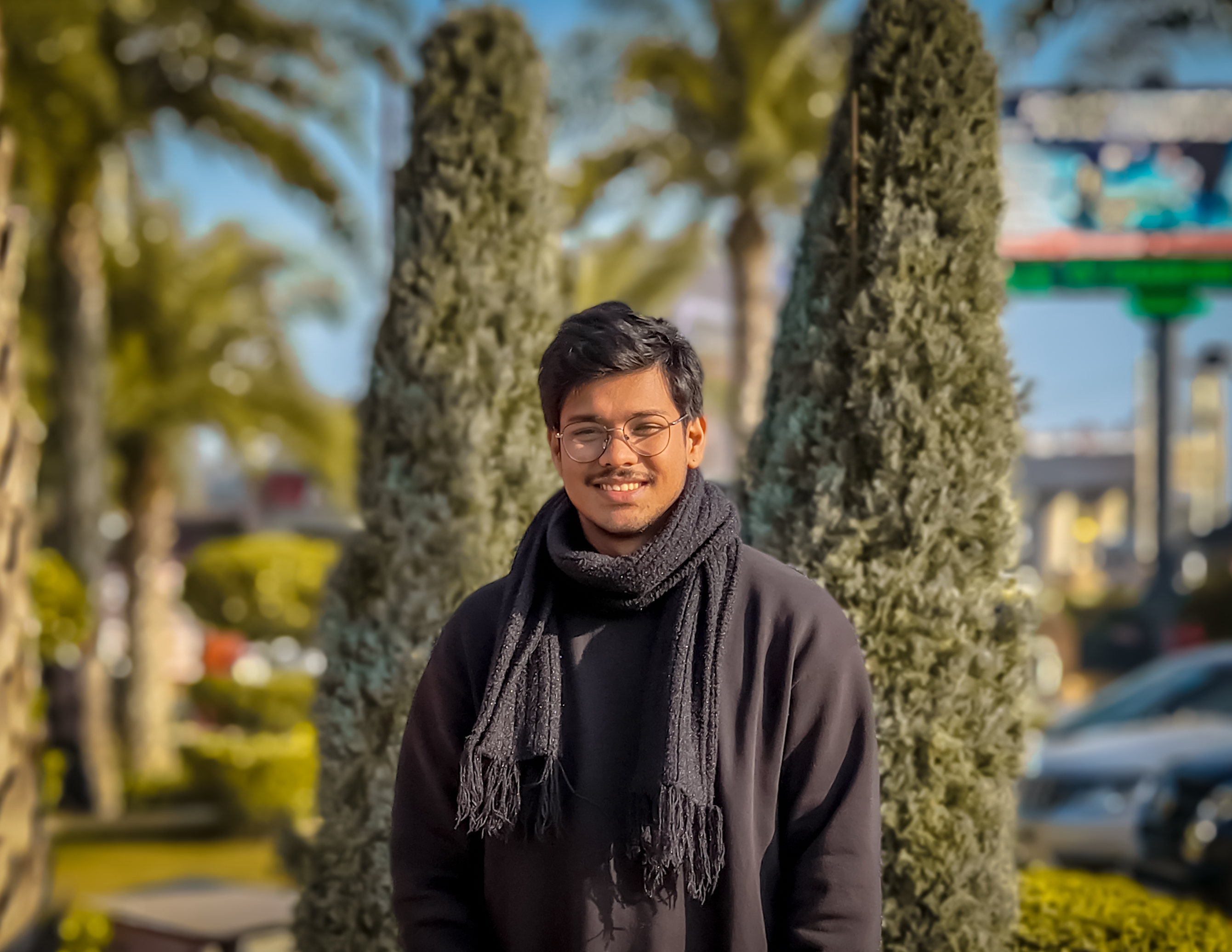 A young man wearing glasses, a black sweater, and a dark scarf is smiling while standing outdoors.