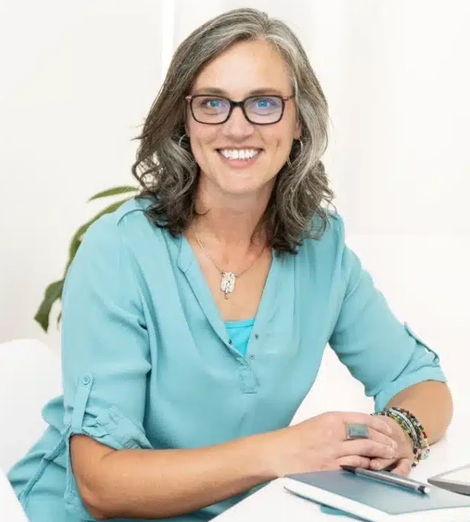 Steph sitting in a white office with a teal shirt, hands folded on desk, and a big smile.