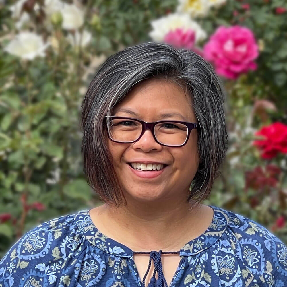 Rosalinda is an asian female with chin-length silver and black hair. She wears glasses and a blue top with a blue and beige print.